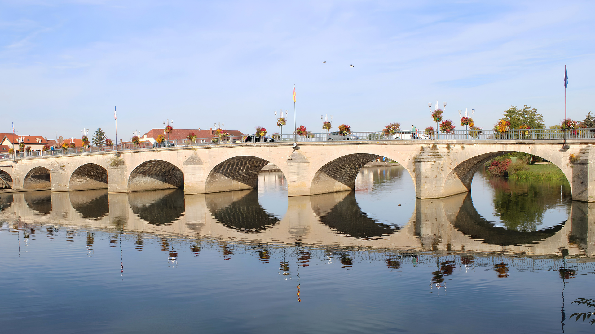 Le pont de pierre de Gray et ses jardinières fleuries, dont les arches se reflètent dans la Saône par un matin calme, en Haute-Saône.
